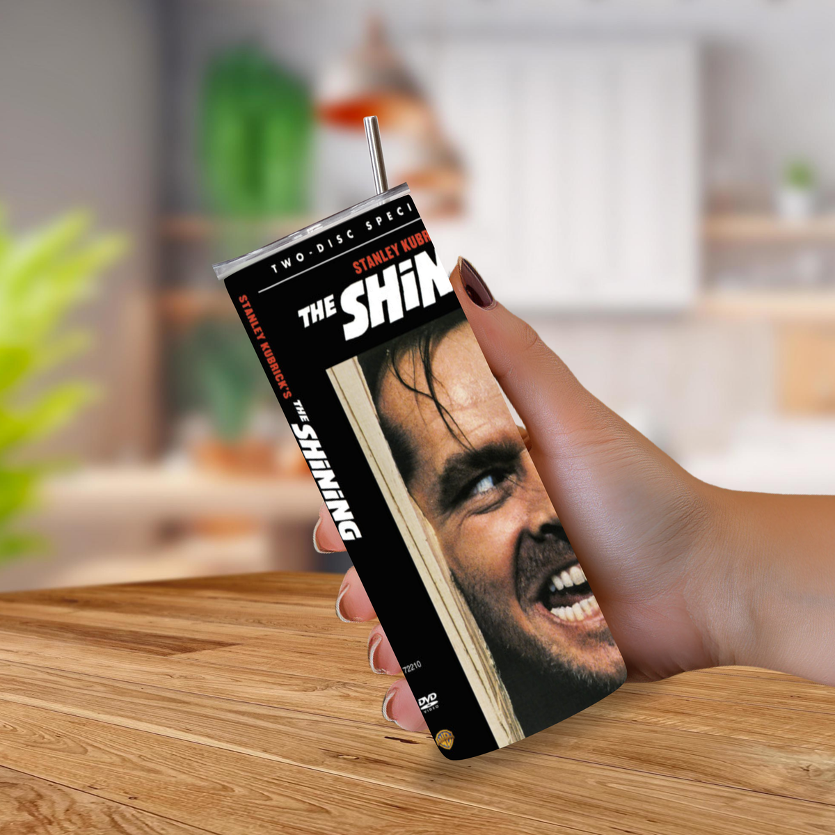 A hand holding a 20oz tumbler designed to look like a DVD case, featuring the movie 'The Shining' cover art, on a wooden table with a blurred kitchen background.