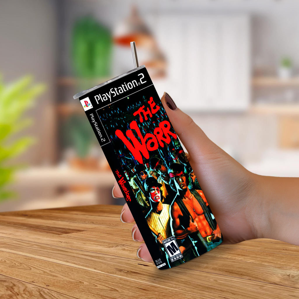 A hand holding a 20oz tumbler designed to look like the PlayStation 2 game case for the video game 'The Warriors'. The device features a printed cover with the PlayStation 2 logo and artwork for 'The Warriors' on a wooden table with a blurred kitchen background.