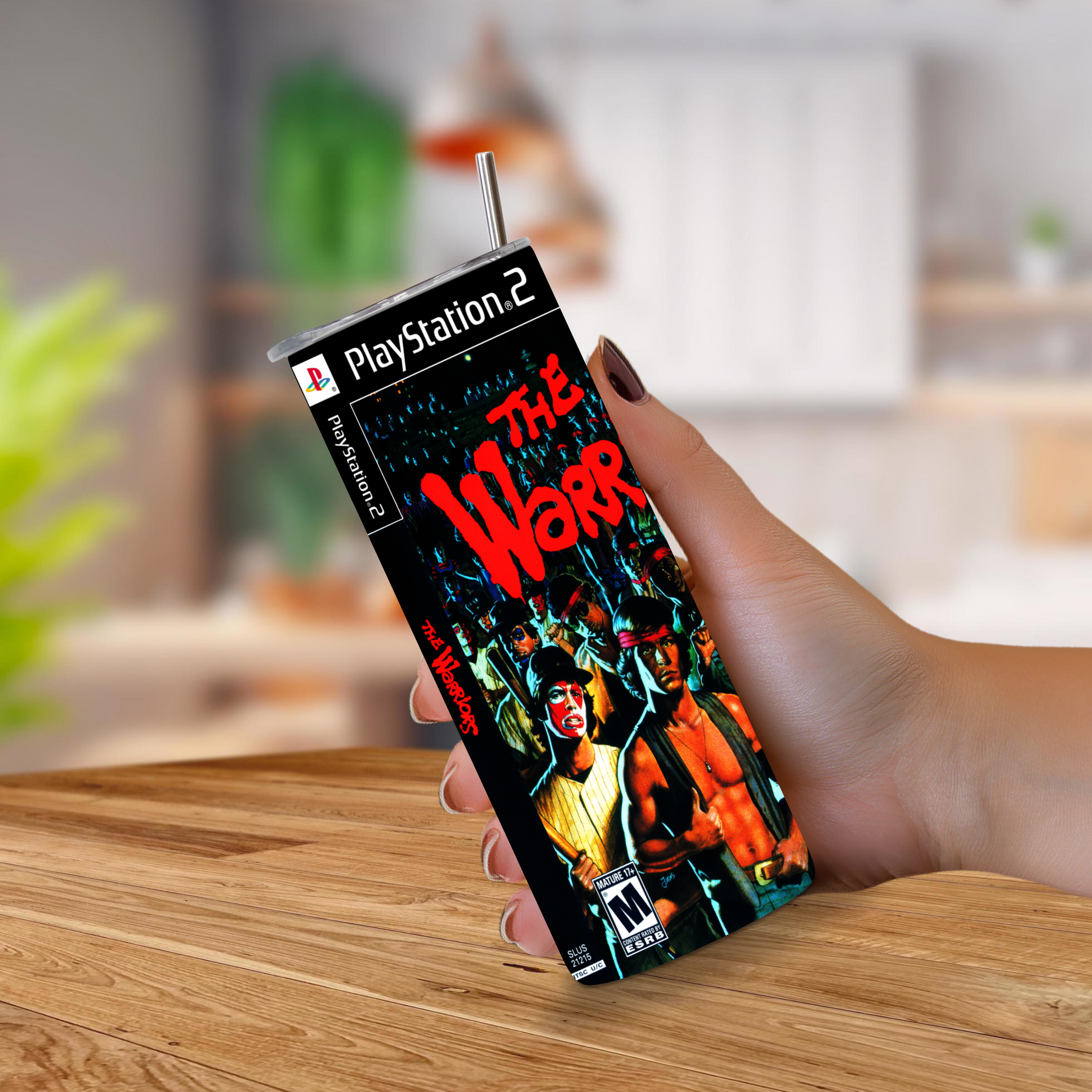 A hand holding a 20oz tumbler designed to look like the PlayStation 2 game case for the video game 'The Warriors'. The device features a printed cover with the PlayStation 2 logo and artwork for 'The Warriors' on a wooden table with a blurred kitchen background.