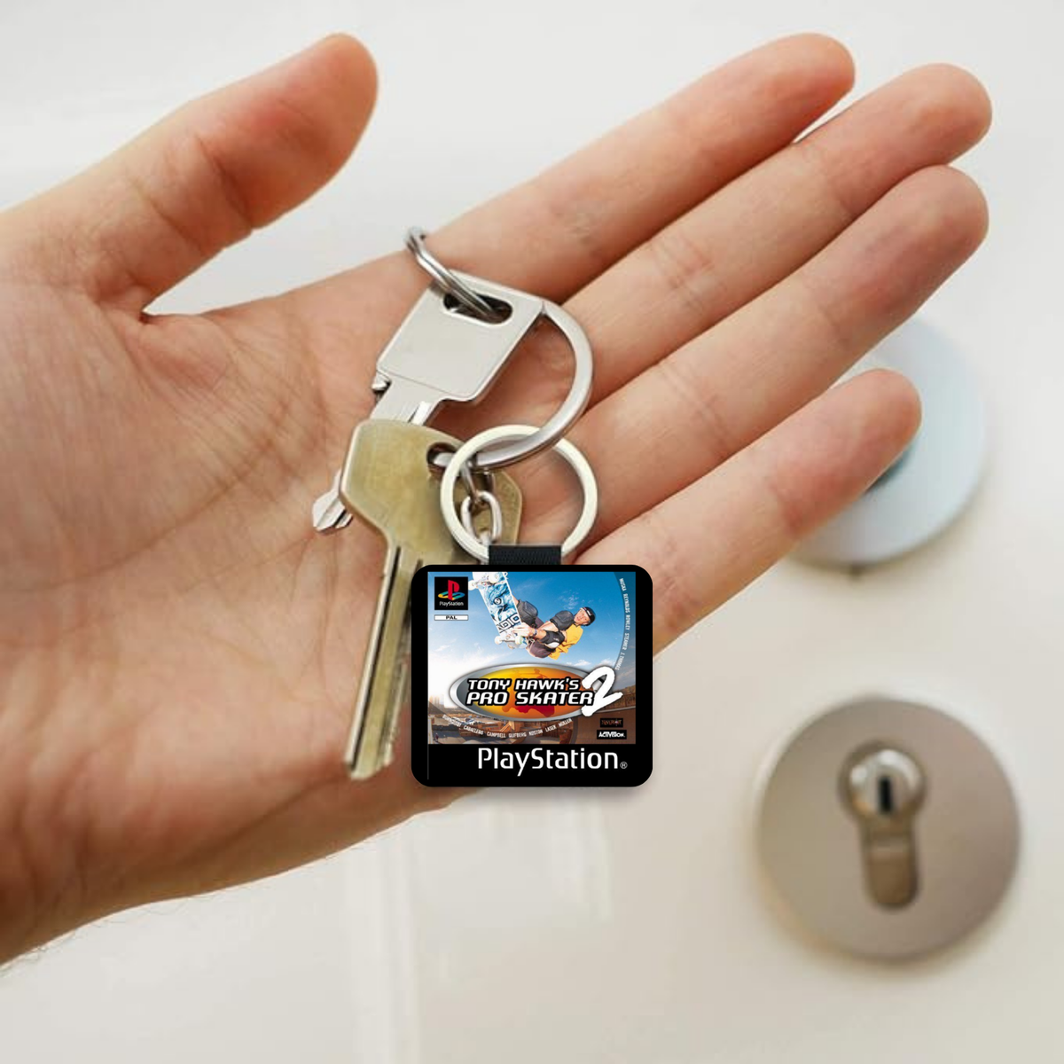 A hand holding a keyring with a small PlayStation-themed Tony Hawk Pro Skater 2 keychain charm and two metal keys. The charm features the PlayStation logo and text, with a blurred lock and keyhole in the background.