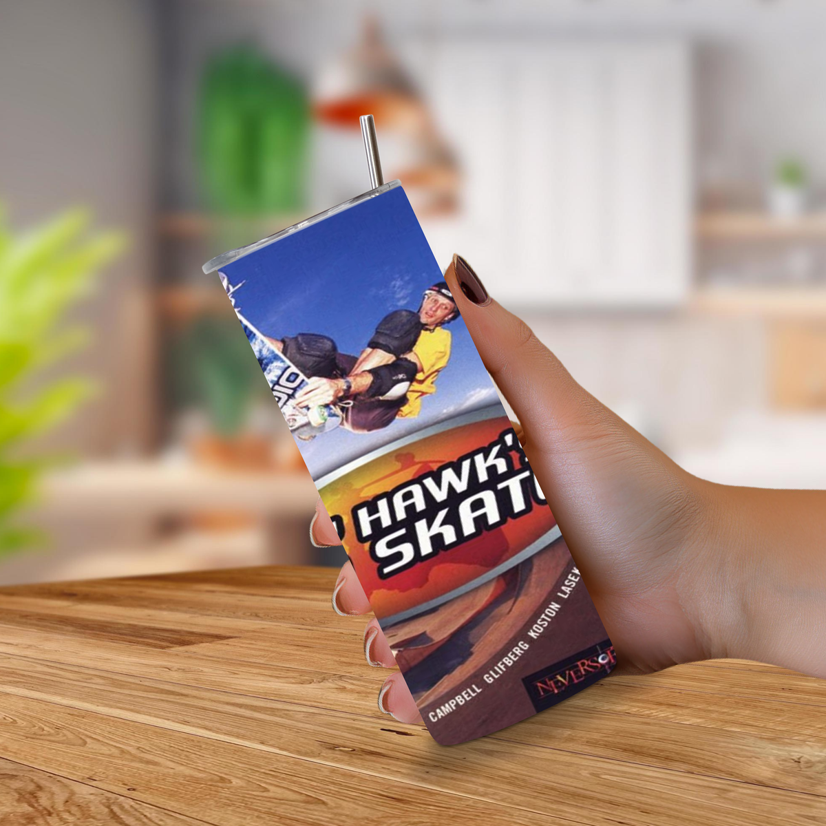 A hand holding a custom 20oz tumbler with a metal straw, featuring a graphic of a snowboarder and the text 'TONY HAWK PRO SKATER 2'. The background shows a blurred indoor environment with a wooden table in the foreground.