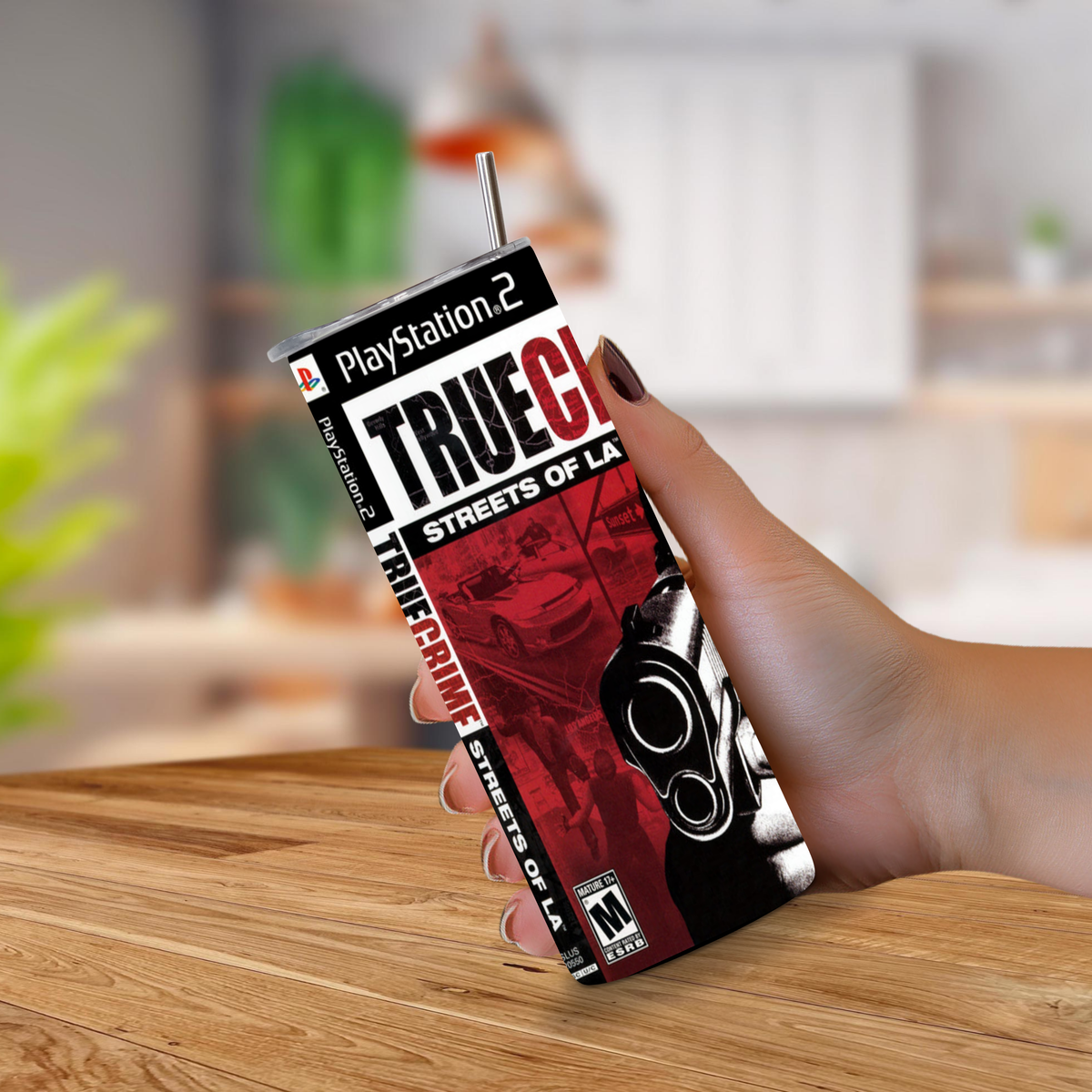 A hand holding a 20oz tumbler featuring PlayStation 2 True Crime: Streets of LA artwork, with a straw inserted at the top, set against a blurred indoor background and wooden tabletop.