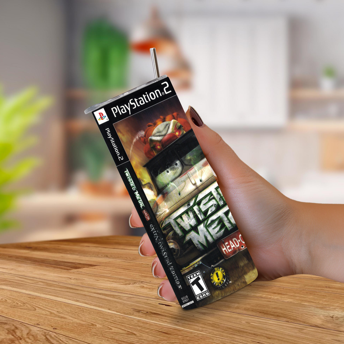 A hand holds a PlayStation 2 game cover styled of a 20oz stainless steel tumbler with a straw, featuring the 'Twist Metal Head On' cover art, against a blurred kitchen background.