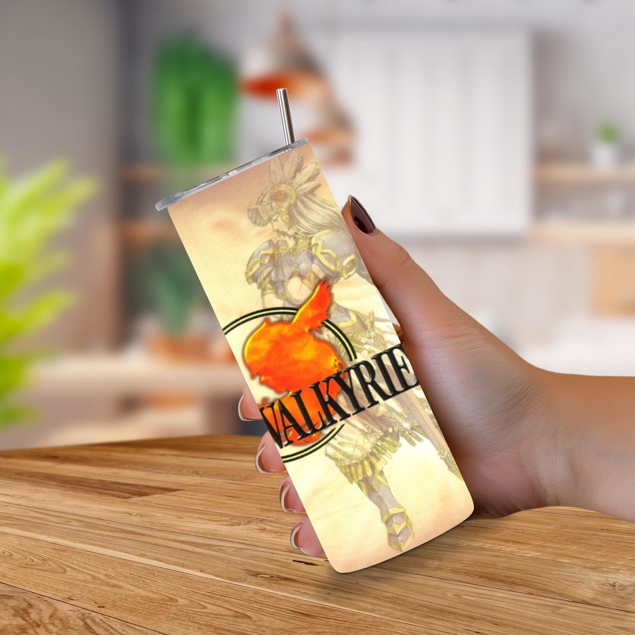 A hand holding a slim 20oz insulated tumbler with a straw, featuring a printed fantasy warrior design and the word 'VALKYRIE PROFILE' on a wooden table with a blurred indoor background.