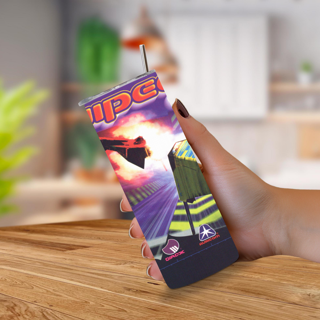 A hand holding a slim 20oz tumbler with a metal straw, featuring a colorful racing graphic and the word 'Wipeout XL' on the front, set against a blurred indoor background with a wooden table.