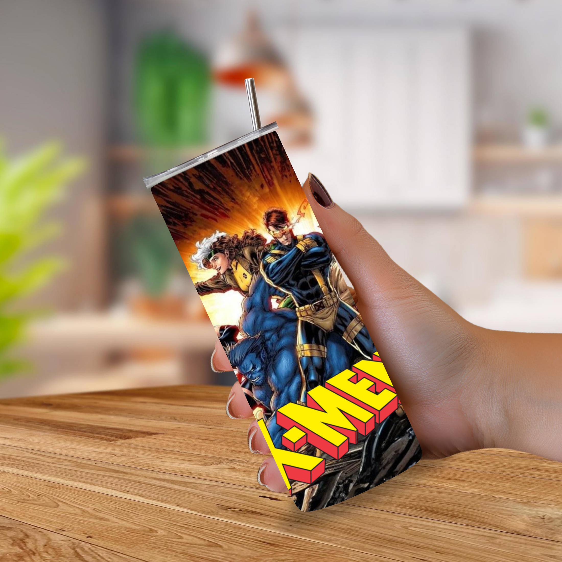 A hand holding a slim rectangular tumbler with a metal straw, featuring a colorful comic-style print of Marvel characters and the large text 'X-Men'. The background is a softly blurred indoor setting with a wooden table visible at the bottom.