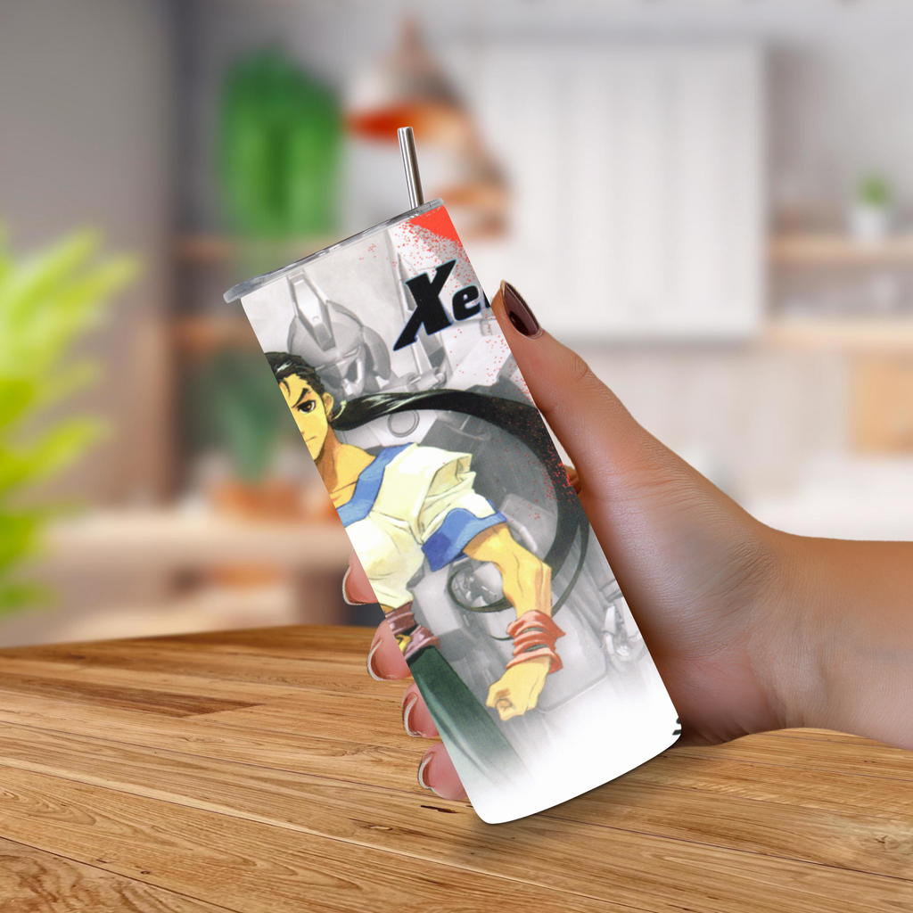 A hand holding a 20oz tumbler with a Xenogears printed anime-style illustration and a metal straw, set against a blurred indoor background with a wooden table.