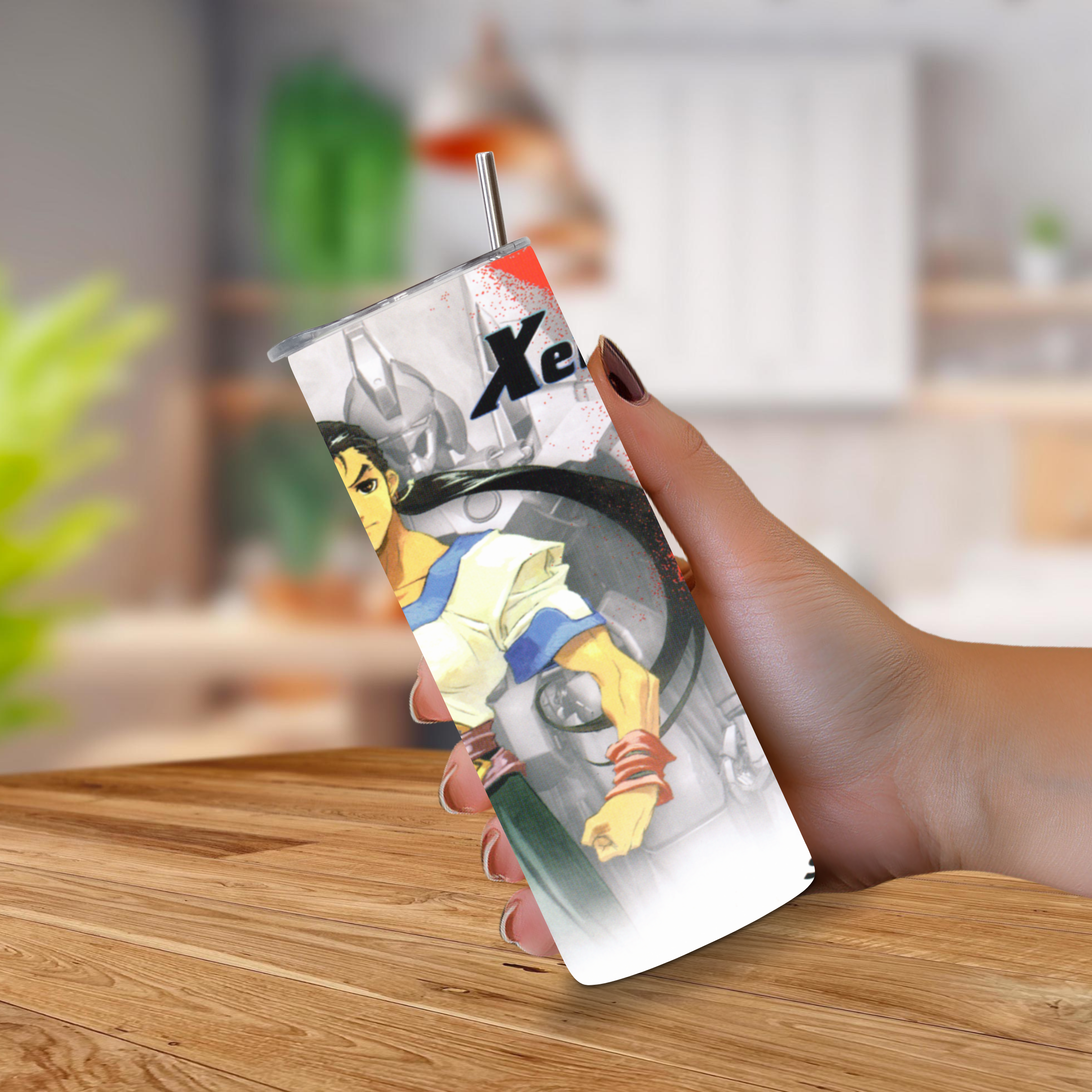 A hand holding a 20oz tumbler with a Xenogears printed anime-style illustration and a metal straw, set against a blurred indoor background with a wooden table.