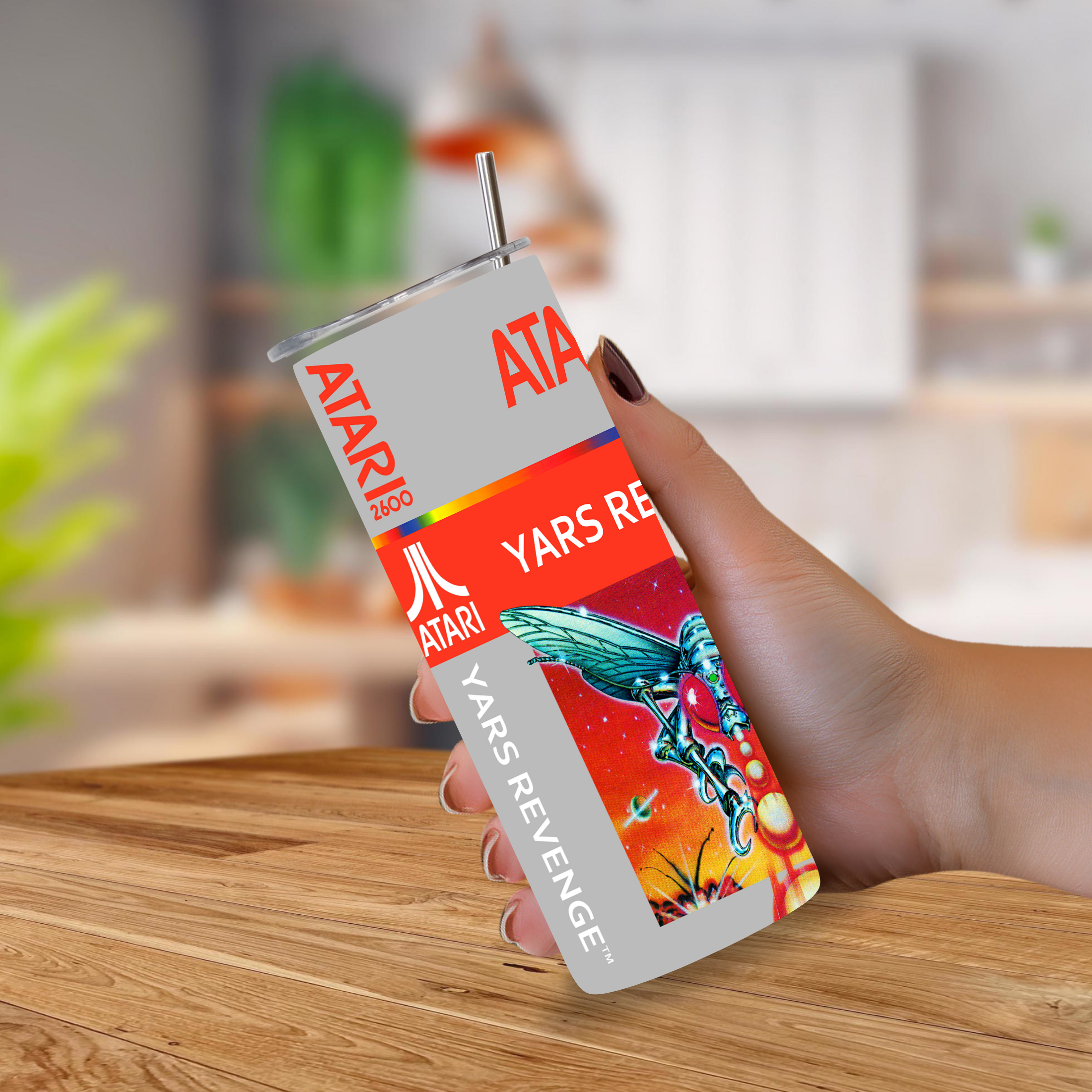 A hand holding a 20oz stainless steel tumbler with a straw, featuring a retro Atari 'Yars Revenge' graphic design, set against a blurred kitchen background.