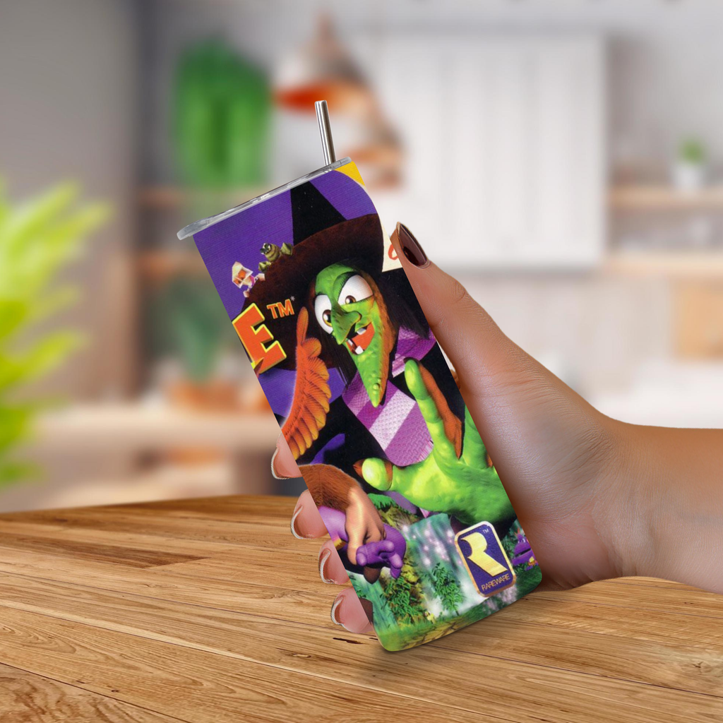 A hand holding a 20oz stainless steel tumbler sleeve with a printed Banjo-Kazooie design, featuring a metal straw, set against a blurred indoor background with a wooden table.