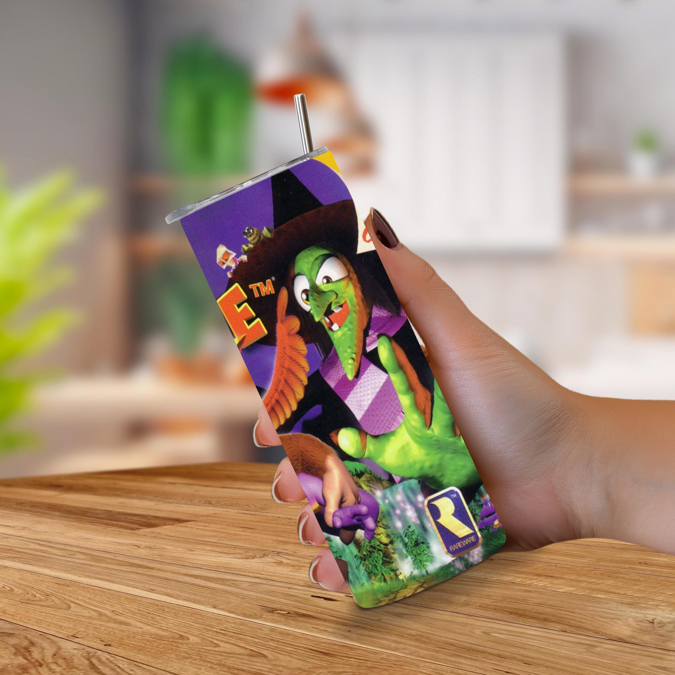 A hand holding a 20oz stainless steel tumbler sleeve with a printed Banjo-Kazooie design, featuring a metal straw, set against a blurred indoor background with a wooden table.