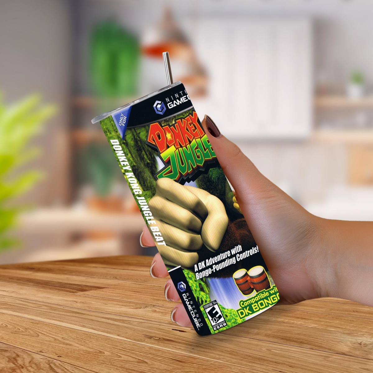 A hand holding a novelty 20oz stainless steel tumbler shaped like Donkey Kong Jungle Beat game cover, with a straw, against a blurred indoor background and wooden tabletop.