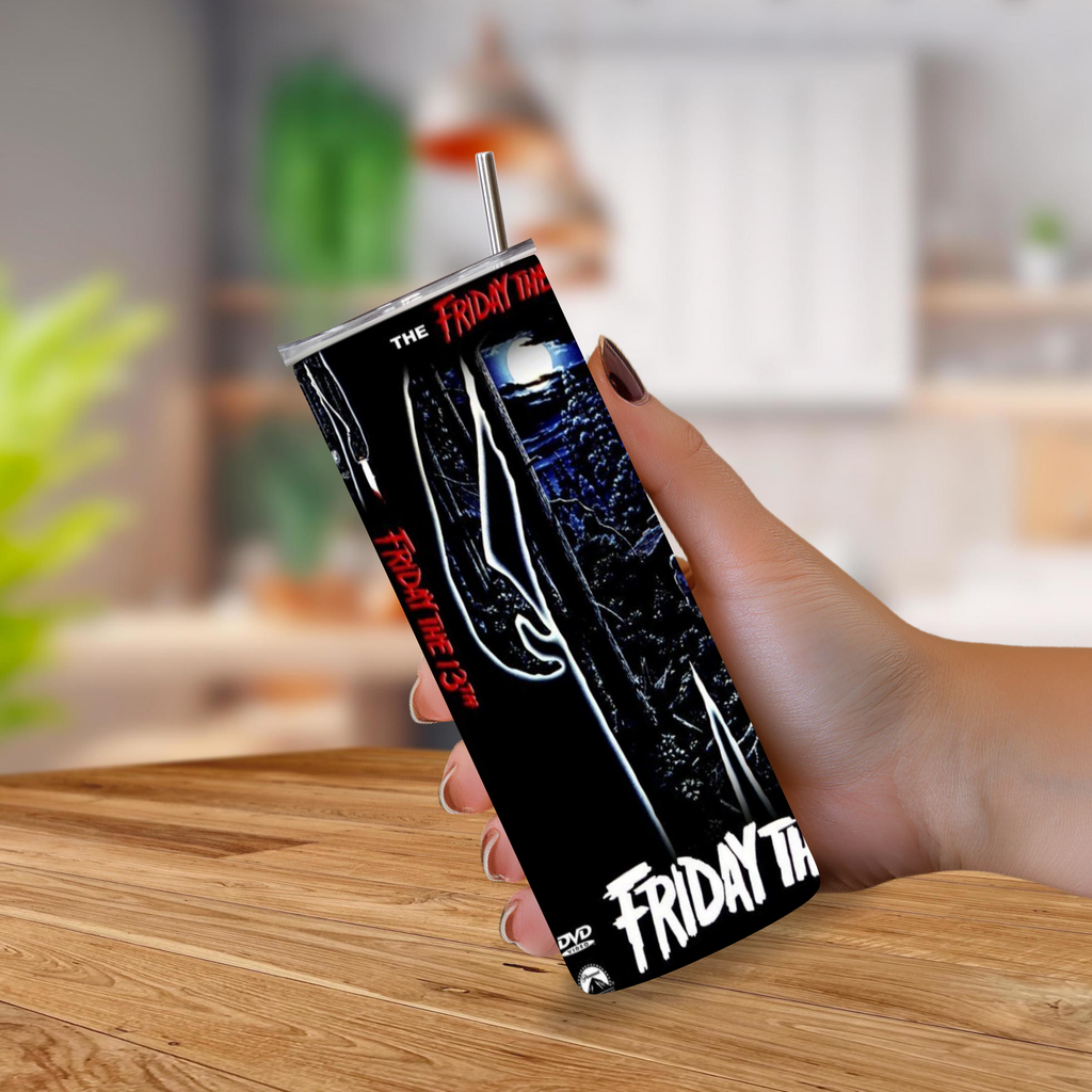 A hand holding a black insulated tumbler with a metal straw, printed with a dark horror-themed graphic and the words 'Friday the 13th'. The background shows a blurred indoor kitchen setting with a wooden table in the foreground.